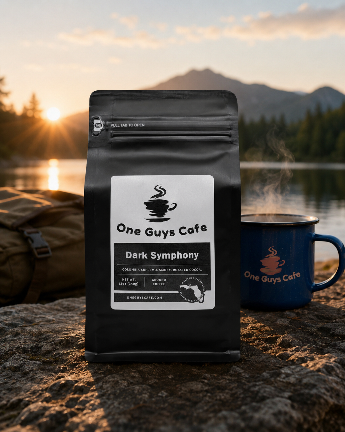  One Guys Cafe Dark Symphony dark roast coffee bag beside a steaming branded mug outdoors at sunrise by a lake.