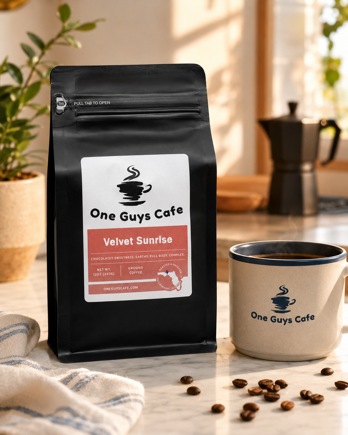One Guys Cafe Velvet Sunrise coffee bag on a bright kitchen counter beside a steaming branded mug.